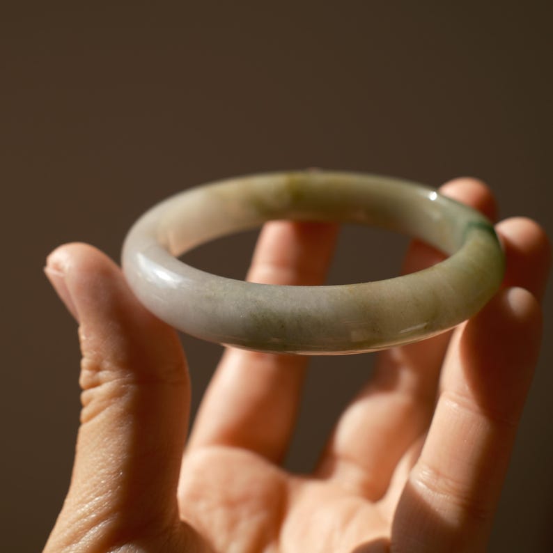 May include: A smooth, circular bangle bracelet crafted from translucent stone with a polished finish. The material displays a variegated appearance, featuring soft gradients of pale grey, off-white, and subtle patches of muted forest green.