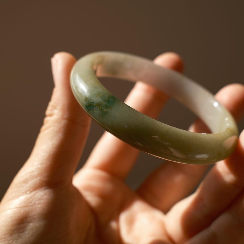 May include: A smooth, polished bangle bracelet crafted from translucent pale green stone. The circular band features subtle variations in color, including soft white tones and a concentrated patch of deep emerald green mineral inclusions.
