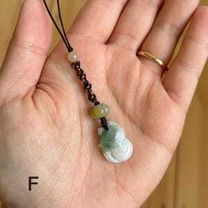 May include: A jade pendant in the shape of a cat, with a black cord. The cat pendant is white and green. The pendant is held in a hand. The letter "F" is in the lower left corner.