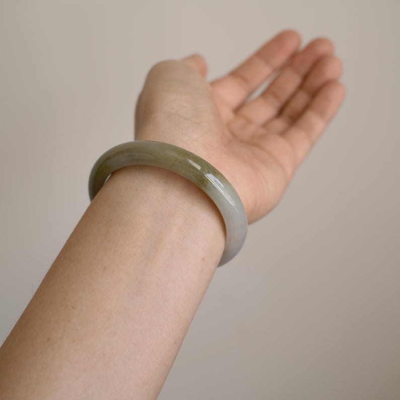 May include: A smooth, polished bangle bracelet crafted from translucent jade stone. The circular accessory features a rounded profile with a natural color gradient transitioning from a soft, pale grey to a warm, muted olive green tone.