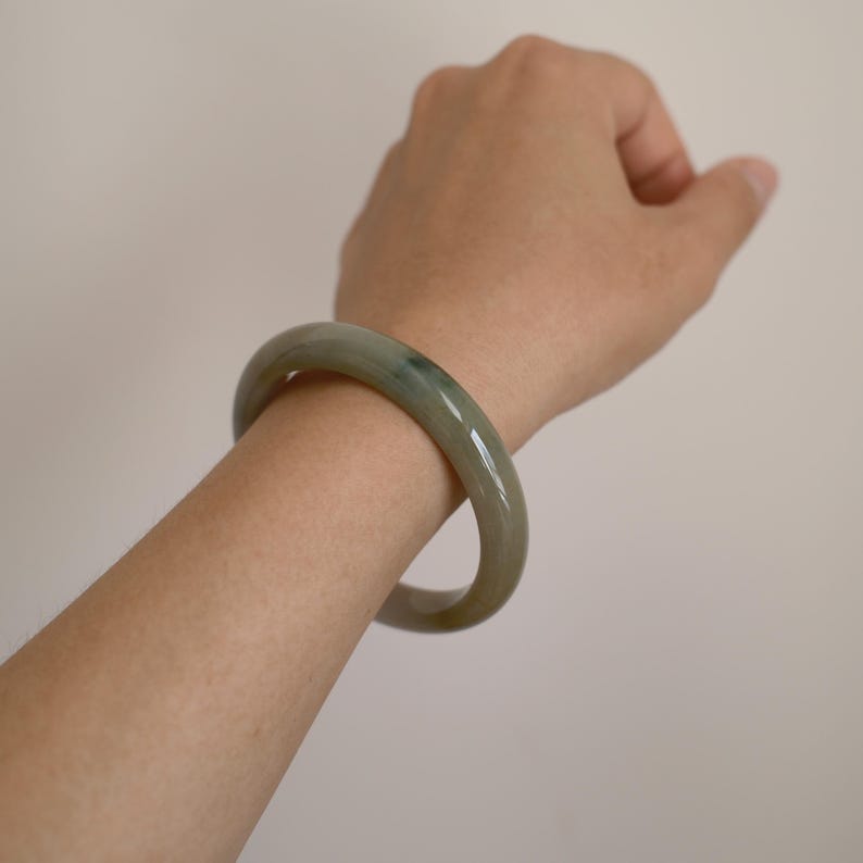 May include: A smooth, polished bangle bracelet crafted from translucent light green stone with subtle darker grey and black inclusions throughout the circular band, featuring a rounded profile and a glossy, reflective surface finish.