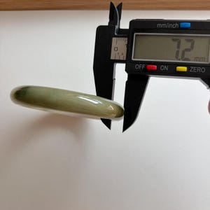 May include: A smooth, rounded, light green jade-colored bangle bracelet being measured by a black digital caliper. The digital display on the caliper clearly shows a measurement of 7.2 mm, indicating the thickness of the bangle's band.