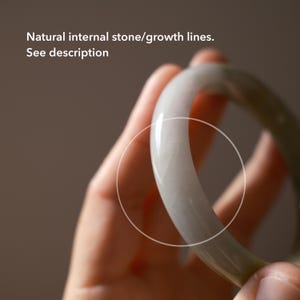 May include: A smooth, pale greyish-green stone bangle bracelet with a polished finish. The surface displays natural internal stone and growth lines. Text on the image reads: Natural internal stone/growth lines. See description.