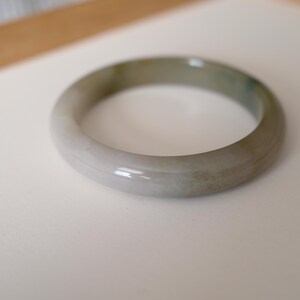 May include: A smooth, polished bangle bracelet crafted from translucent stone with a rounded profile. The material displays a variegated appearance, featuring soft gradients of pale grey, off-white, and subtle hints of muted green throughout the circular band.