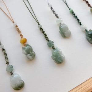 May include: Close-up of several jade fox pendants with various cord colors. The pendants are carved in a sitting fox shape, with shades of green and white. The cords are in shades of brown, green, and gray, with decorative beads.