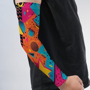 May include: An arm sleeve featuring a vibrant, multi-colored geometric pattern with bright orange, hot pink, yellow, and teal shapes. The design includes various abstract elements such as circles, triangles, and lines scattered across the fabric.