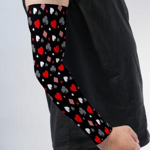 May include: A black arm sleeve featuring a repeating pattern of red hearts, white spades, grey clubs, and pink diamonds. The fabric is form-fitting and covers the forearm and bicep area, displaying the four classic playing card suit symbols.