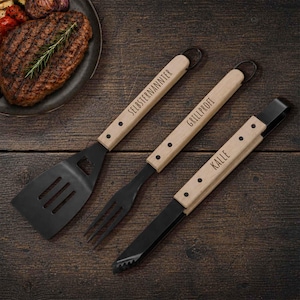 Op de afbeelding: Een set van drie grillgereedschappen met houten handvatten en zwarte metalen koppen. De gereedschappen omvatten een spatel, een vork en een tang. De handvatten hebben de woorden "SELBSTERNANNTER", "GRILLPROFI" en "KALLE".
