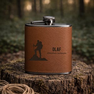 Könnte beinhalten: Eine braune, lederbezogene Flachmann mit einem Edelstahl-Deckel. Sie zeigt eine schwarze Silhouette eines Wanderers und den Namen "OLAF" mit dem Satz "draußen zuhause". Die Flasche steht auf einem Baumstumpf.