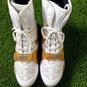 May include: Pair of white and gold Nike wrestling shoes. The shoes have a high-top design with a mesh and leather upper. Gold accents are visible on the sides and straps, with the Nike logo prominently displayed. White laces and a gold strap with the Nike logo complete the design.