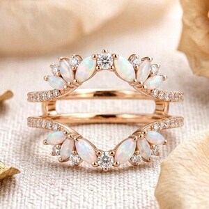 Op de afbeelding: Een roségouden ringbeschermer met twee gebogen banden die aan de basis zijn verbonden. Het ontwerp is versierd met marquise-geslepen opalen en kleine ronde briljant geslepen diamanten in een symmetrisch, bladachtig patroon langs de randen.