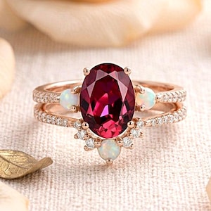 Könnte beinhalten: Ein Ring aus Roségold mit einem großen, oval geschliffenen, tiefroten Granat als Mittelstein, akzentuiert durch drei kleine, runde weiße Opale und eine Ringschiene, die mit winzigen, klaren Pavé-Diamanten in einem filigranen Design besetzt ist.