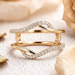 May include: A gold ring guard featuring two wavy bands connected at the sides. Both bands are encrusted with small, clear, sparkling stones along their top edges, creating a delicate, layered appearance with a polished metallic finish.