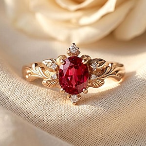Può includere: Un anello in oro con una pietra preziosa rossa centrale a taglio ovale circondata da piccole pietre chiare. La fascia presenta dettagliate incisioni a forma di foglia che si estendono dalla montatura centrale in metallo.