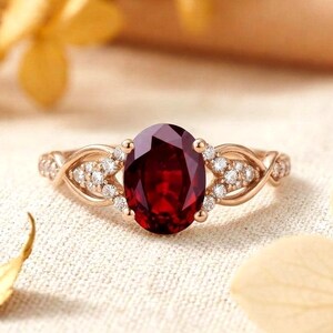 Könnte beinhalten: Ein Ring aus Roségold mit einem großen, oval geschliffenen, tiefroten Edelstein in der Mitte. Die Ringschiene ist auf beiden Seiten mit einem komplizierten, gedrehten Metallmuster gestaltet, das durch kleine, runde, klare Steine akzentuiert wird.