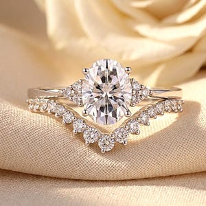 Peut inclure: Une bague de fiançailles argentée avec une pierre centrale ovale transparente entourée de petites pierres rondes, accompagnée d'une alliance en forme de chevron ornée d'une rangée de petites pierres rondes transparentes.