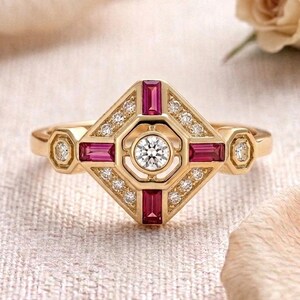 May include: A gold ring featuring a central round brilliant-cut diamond surrounded by four rectangular baguette-cut rubies arranged in a geometric diamond shape, accented with small round diamonds along the frame and on the side shoulders.