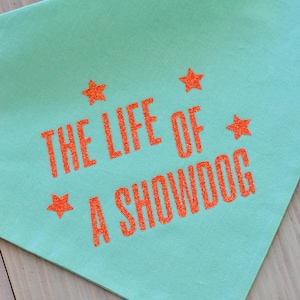May include: A light teal dog bandana with the text "THE LIFE OF A SHOWDOG" in orange glitter lettering. The design includes orange glitter stars. The bandana is made of soft fabric.