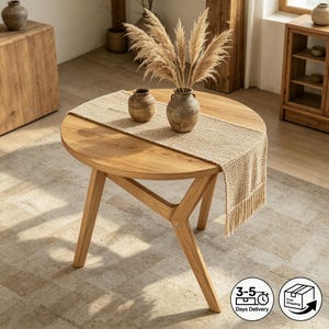 May include: A round wooden coffee table with a natural finish. A woven beige table runner with fringe detail is centered on the table. Two small ceramic vases and a larger vase with dried pampas grass are on the table.