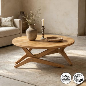 May include: A round, light oak coffee table with a crossed-leg base. The table is adorned with a vase of dried flowers, a candle in a holder, and a woven tray. The table is set on a neutral-toned rug.