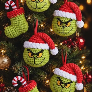 May include: A collection of handmade crochet Christmas ornaments featuring green character heads with yellow eyes and red Santa hats, alongside green crochet stockings containing red and white striped candy canes, all hanging by red ribbons.