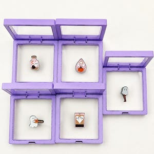 May include: Five square lavender floating display frames with clear windows, each containing a unique enamel pin. The pins feature a ghost in a bottle, a pumpkin on a planchette, a ghost with a chainsaw, a tarot card, and a ghost with legs.