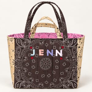 May include: A quilted tote bag featuring a dark brown, tan, and pink paisley bandana print. The front displays the name JENN in pastel-colored embroidered letters, accented by four small red hearts. It has two sets of handles in brown and tan.