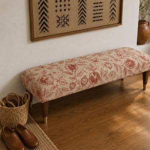 May include: A rectangular bench with a beige fabric featuring a red pomegranate pattern. The bench has four wooden legs with gold-coloured tips. The bench is placed on a wooden floor, with a woven basket and shoes nearby.