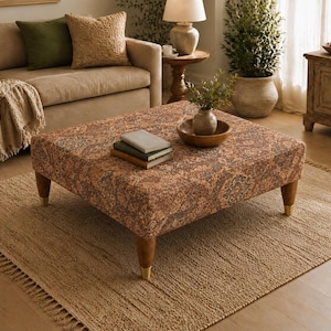 May include: A square ottoman featuring a multi-coloured, intricate paisley-inspired pattern in shades of rust, brown, and muted blue. The piece is supported by four tapered wooden legs with metallic gold-toned caps at the base of each leg.