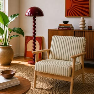 May include: A living room scene featuring a striped armchair, a round wooden coffee table, and a burgundy floor lamp with a spiral stem. A wooden cabinet and artwork are also visible, creating a cozy, inviting atmosphere.