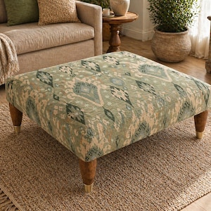 Puede incluir: Un otomán cuadrado con tela estampada en tonos verdes, beige y azul. Tiene patas de madera con puntas doradas y descansa sobre una alfombra de yute tejida. El diseño combina elementos geométricos y florales. Dimensiones: aproximadamente 90 cm x 90 cm.