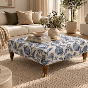Op de afbeelding: Een vierkante poef bekleed met een witte stof met een blauwe botanische print van granaatappels en bladeren. Het meubelstuk rust op vier taps toelopende houten poten met goudkleurige metalen doppen aan de onderkant voor een stabiele basis.