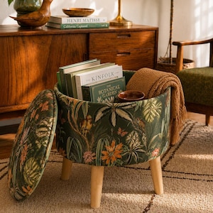 May include: A round storage ottoman with a botanical print in shades of green, orange, and pink. The ottoman has wooden legs and is filled with books and a brown blanket. The lid is open, revealing the interior.