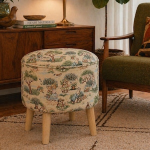 May include: A round upholstered footstool with a whimsical pattern featuring rabbits, bears, trees, and floral elements in shades of beige, green, and blue. The stool has four wooden legs.