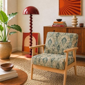 May include: A living room scene featuring a light wood armchair with a patterned green and blue cushion. A red floor lamp with a spiral base stands nearby. A wooden cabinet and artwork complete the cozy interior.