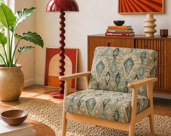 Könnte beinhalten: Wohnzimmer-Interieur mit einem hellen Holzarmstuhl mit einem gemusterten grün-blauen Kissen. Eine rote Stehlampe mit spiralförmigem Fuß ist zu sehen. Ein Holzschrank und Kunstwerke vervollständigen die Szene.