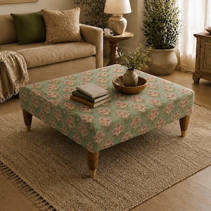 May include: A square ottoman upholstered in a sage green fabric featuring a repeating pink and white floral pattern. The piece is supported by four tapered wooden legs with gold-toned metal caps at the base, providing a stable foundation.