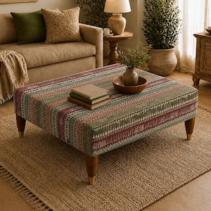 May include: A square ottoman featuring a multi-colored striped fabric pattern in shades of green, red, and cream. The piece is supported by four tapered wooden legs with gold-toned metal caps at the base, providing a stable foundation.