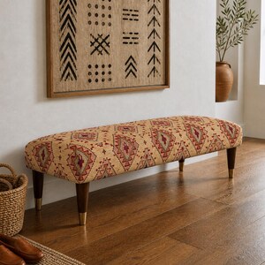 Puede incluir: Banqueta tapizada con un patrón geométrico beige y rojo. La banqueta tiene patas de madera oscura con puntas color latón. Una cesta tejida y una obra de arte enmarcada están en el fondo. Es un mueble decorativo.
