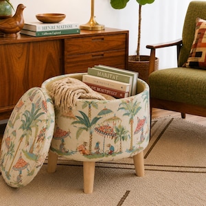 May include: An open, round ottoman with a lid, featuring a tropical print of palm trees, umbrellas, and figures. The ottoman has light wood legs and is filled with books and a beige throw blanket. The lid is placed to the side.