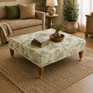 Peut inclure: Un pouf carré recouvert d'un tissu crème orné d'un motif toile de jouy vert représentant des scènes pastorales. Le meuble repose sur quatre pieds en bois tourné avec des embouts métalliques dorés, offrant une base stable au plateau rembourré.