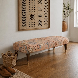 May include: An upholstered bench with a floral pattern in shades of peach, blue, and red. The bench has dark wooden legs with gold-colored tips. A woven basket and a pair of brown leather shoes are in the foreground.