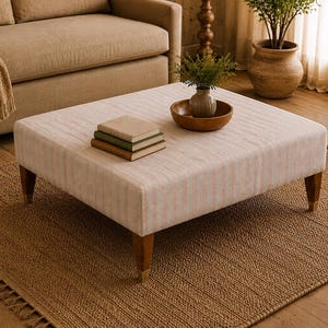 May include: A square ottoman upholstered in a light beige fabric with subtle vertical stripes, featuring four tapered wooden legs with metallic feet. The top surface holds a stack of three books and a wooden bowl containing a small green plant.