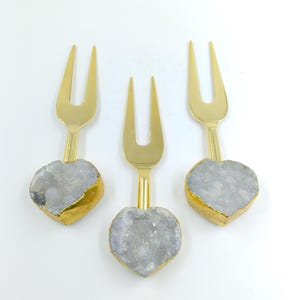 Puede incluir: Tres tenedores dorados para servir con mangos de piedra gris y blanca en forma de corazón. Los bordes de los mangos en forma de corazón están ribeteados en dorado. Los tenedores están dispuestos sobre un fondo blanco.