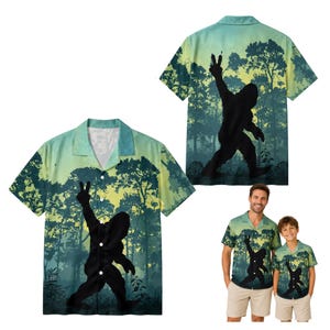 May include: A short-sleeve button-down shirt featuring a forest landscape print in shades of green and yellow. The design displays a black silhouette of a creature making a peace sign gesture. The shirt has a notched collar and a front button closure.