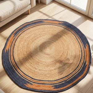 May include: A round jute rug with a natural tan center and a border of navy blue and orange geometric patterns. The rug is placed on a light wood floor, near a beige sofa and a potted plant.