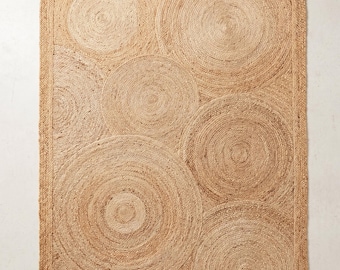 May include: A rectangular area rug crafted from natural tan woven fibers, featuring a series of overlapping circular patterns throughout the surface. The design is bordered by a straight, braided edge that frames the circular motifs.