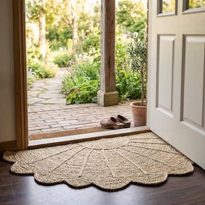May include: A semi-circular natural fibre doormat with a scalloped edge and a sunburst pattern of woven rope. The mat is placed on a dark wooden floor inside a doorway that opens to a brick patio, a potted plant, and a pair of brown clogs.