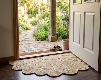May include: A semi-circular natural fiber doormat with a scalloped edge and a sunburst pattern of woven rope. The mat is placed on a dark wooden floor inside a doorway that opens to a brick patio, a potted plant, and a pair of brown clogs.