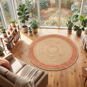 May include: A large, round, woven area rug featuring a natural tan center with concentric rings in shades of orange, pink, and red. The rug is positioned on a light-colored wooden floor in a room filled with various potted green plants.